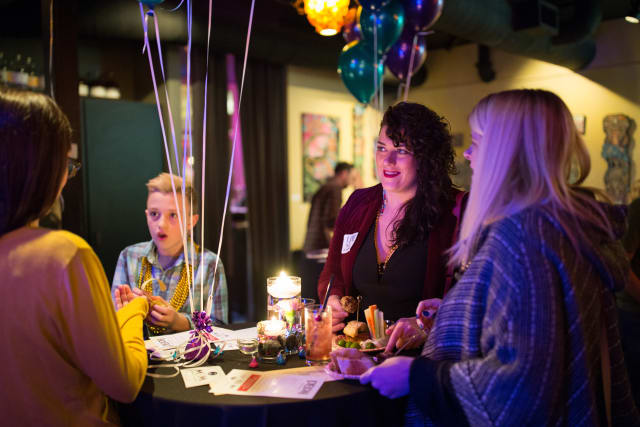 Group of attendees engaging at a table during the Love Bomb 2017 event, featuring colorful balloons, food, and drinks, highlighting community support for My Voice Music.